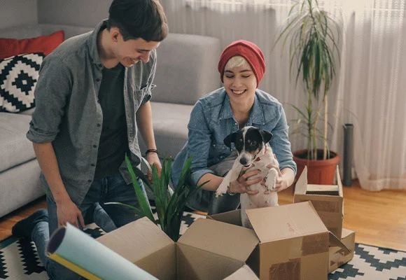 Small squared image of a couple unboxing as they move into their new home, with woman holding dog