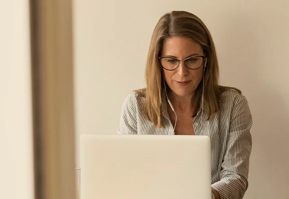 Squared image of woman sat at laptop computer looking at screen and listening through headphones.