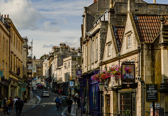 Image from the bottom of a city centre street in Bath, looking uphill. Pubs, shops and other commercial properties line the street with cards and people heading up and down the street.